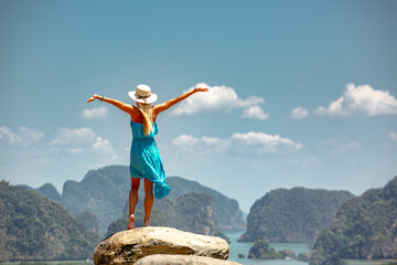 Beautiful young lady in blue dress and white hat is standing with open arms on big rock with sea and islands view. Phang Nga province, Thailand - obrazy, fototapety, plakaty