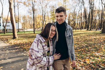 Heterosexual caucasian young loving couple walking outside in the city park in sunny weather, hugging smiling kissing laughing spending time together. Autumn, fall season, orange yellow red maple leav