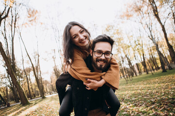 Heterosexual caucasian young loving couple, husband carrying on back his wife in sunny weather, hugging smiling kissing laughing spending time together. Autumn, fall season, orange yellow red maple le - obrazy, fototapety, plakaty