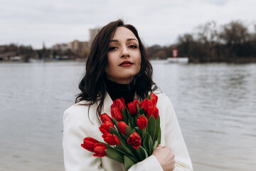 Attractive brunette woman walking on the beach shore in moody cloudy windy weather with bouquet of red tulips flowers, dressed in white suit jacket. International Women`s Day 8th March concept
