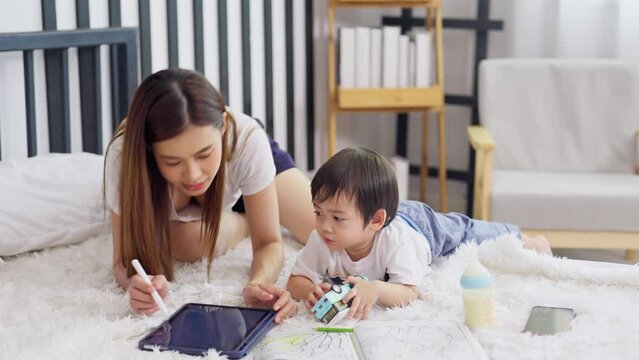 Asian mother sit on bed using tablet to enjoy with little son who paint on child book and they stay on bed together.