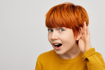 Red-haired boy boy holds hand near the ear listening to sounds and voices, on grey background....