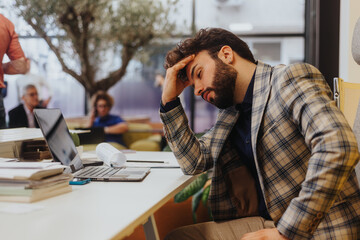 Young, male, business person struggling with his tasks at work. He is holding his forehead while having a headache.