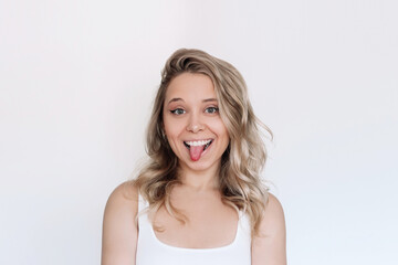 A young attractive caucasian smiling blonde woman with wavy hair showing tongue grimacing with a funny face isolated on a white background - obrazy, fototapety, plakaty