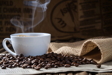 White cup of hot coffee with steam over scattered roasted beans and burlap