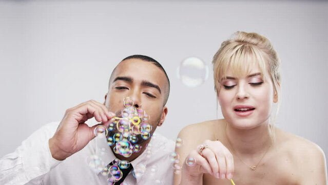 Face, wedding and interracial couple blowing bubbles in studio on white background for celebration of love. Party, event or photobooth with young man and woman together for marriage or bonding