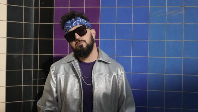 Stylish male in a shiny silver jacket and sunglasses exudes confidence with a cool demeanor against a vibrant tiled wall.