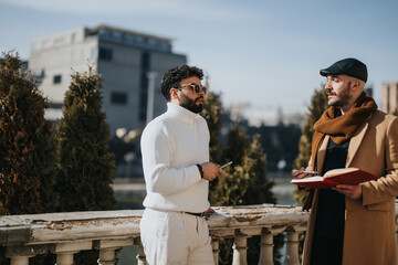 Naklejka premium Fashionably dressed men having a serious discussion on a balcony with cityscape in the background. Warm sunlight enhances the friendly atmosphere.