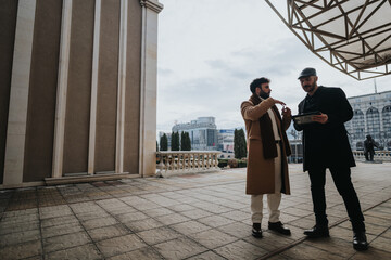 Two well-dressed men engaged in a serious conversation while holding a tablet. The setting displays elegant classical columns and urban surroundings.
