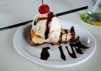 Slice of cheesecake topped with a scope of ice cream, cherry and splattered with chocolate.