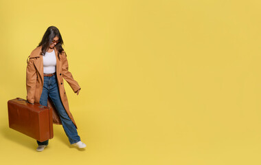 a woman in jeans, a white T-shirt, and a brown coat carrying a suitcase and taking a selfie by phone on a yellow background. Happy people going on holiday, vacation