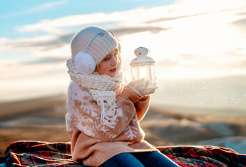 Teenager girl in Christmas sweater sitting on top of a mountain  with lantern with magical light on...