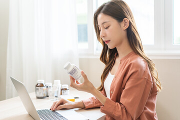 Wellness and dieting asian young woman, girl working from home using computer, typing or searching prescription on medicine label about vitamins information online, holding bottle of food supplement. - obrazy, fototapety, plakaty