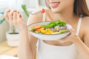 Deurstickers Diet concept, happy asian young woman hand use a fork to prick tomato, fresh vegetable or green salad, eat nutrition food  on table at home, low fat to good body. Girl getting weight loss for healthy. © KMPZZZ
