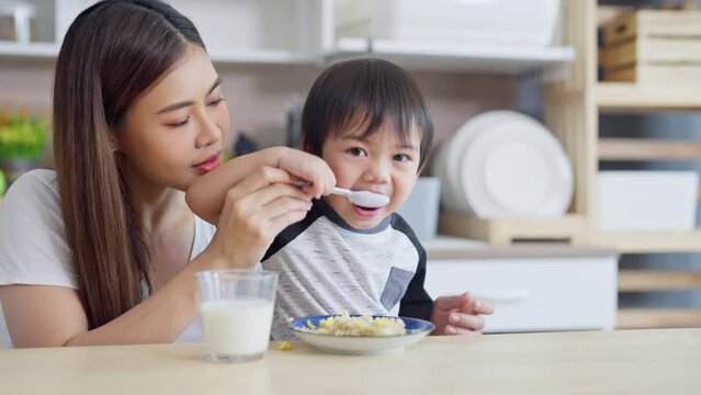 Young Asian mother support little boy to eat food in kitchen by help to suport hold spoon for the child.