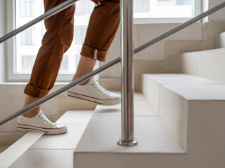 Woman in white sneakers and khaki trousers goes upstairs to her apartment. White staircase in apartment building. Casual outfit, urban fashion. Physical exercises. - 721464608