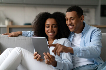 Happy black couple using digital tablet while relaxing on sofa in living room, shopping online,...