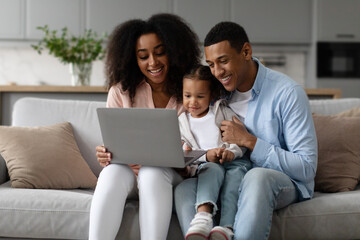 Happy African American parents and their little daughter using laptop at home, watching movie, browsing internet, sitting on couch in living room