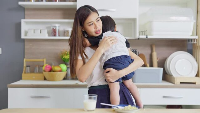 Wide shot of young Asian woman carry little boy lean to her shoulder during eat food in the kitchen. 