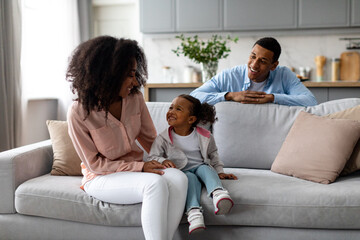 Happy black parents resting with cute little daughter, sitting on sofa in living room interior,...