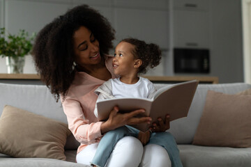 Loving black mother reading book with her little daughter, sitting together on sofa at home,...