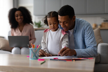 Loving black father teaching little daughter drawing while mother working on laptop on background,...