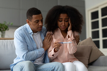 Upset black couple looking at negative pregnancy test sitting together on couch at home, caring...