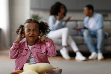Black baby girl covering ears while her parents arguing on background, depressed child feeling lonely in living room at home - obrazy, fototapety, plakaty