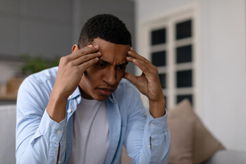 Sad depressed black man sitting on sofa touching head, suffering from headache, migraine, having...
