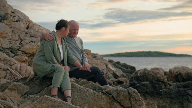 charming mature spouses enjoying vacation by the sea together sitting on shore backdrop of sunset