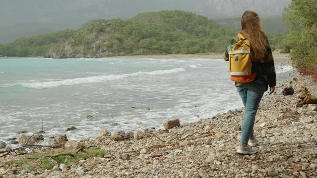 A woman travel blogger walks along the rocky shore of the sea against the backdrop of mountains in the clouds, capturing moments with photos for social media or as memories.