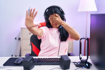 Mature hispanic woman playing video games at home covering eyes with hands and doing stop gesture with sad and fear expression. embarrassed and negative concept.