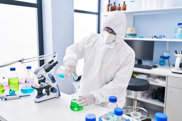 Young hispanic man scientist wearing medical mask pouring liquid on test tube at laboratory