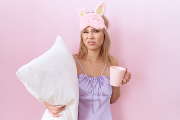 Young caucasian woman wearing pajama hugging pillow and drinking coffee clueless and confused...