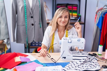 Young blonde woman tailor smiling confident sitting on table at tailor shop