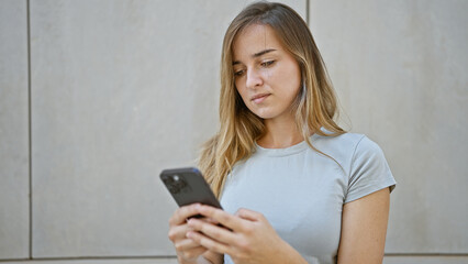 Cool, young blonde woman in casual fashion, seriously engrossed in her smartphone outside on urban...