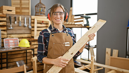 Attractive young blonde woman carpenter with headphones expertly handles wood plank in cozy carpentry workshop