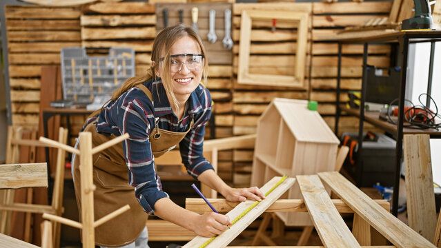 Smiling, beautiful blonde young woman carpenter standing in a workshop, proudly measuring and marking a wood plank inside indoor carpentry studio