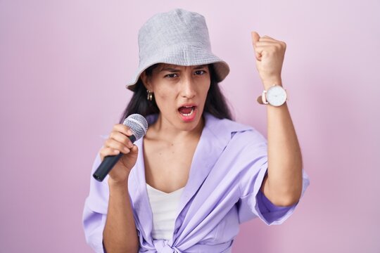Young hispanic woman singing song using microphone over pink background annoyed and frustrated shouting with anger, yelling crazy with anger and hand raised