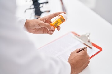 Young hispanic man pharmacist holding pills bottle writing on document at pharmacy
