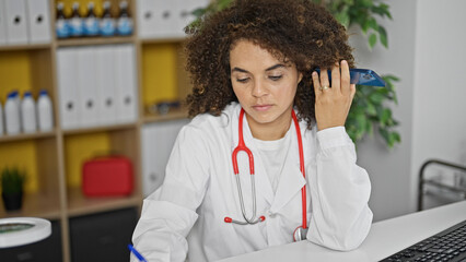 Young beautiful hispanic woman doctor listening to voice message by smartphone writing medical...