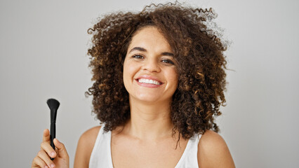 Young beautiful hispanic woman smiling confident holding make up brush over isolated white...