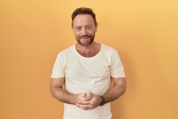 Middle age man with beard standing over yellow background with hands together and crossed fingers smiling relaxed and cheerful. success and optimistic