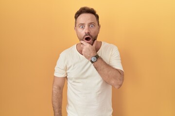 Middle age man with beard standing over yellow background looking fascinated with disbelief, surprise and amazed expression with hands on chin