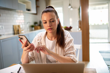 Young female freelancer working on laptop using smartphone at home
