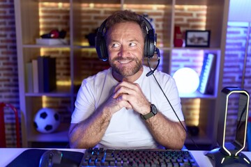 Middle age man with beard playing video games wearing headphones laughing nervous and excited with...