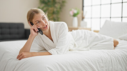 Young blonde woman wearing bathrobe lying on bed talking on smartphone at bedroom