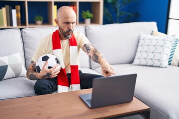 Hispanic man with tattoos watching football match hooligan holding ball on the laptop pointing with...