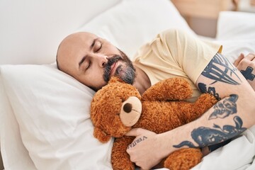 Young bald man hugging teddy bear lying on bed sleeping at bedroom