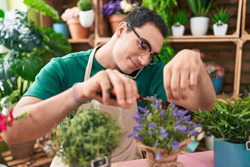 Young hispanic man florist talking on smartphone cutting lavender plant at flower shop - obrazy, fototapety, plakaty